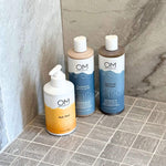 Botanical Bath Trio featuring OM Botanical sulfate-free shampoo, conditioner and styling gel, and body wash on a tiled shower floor. Ideal for gentle cleansing and hydration.