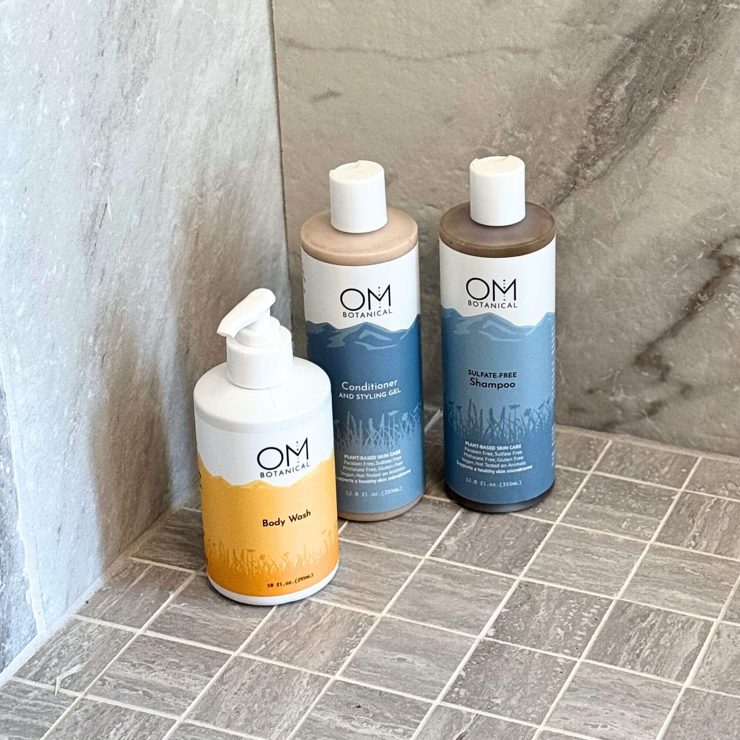 Botanical Bath Trio featuring OM Botanical sulfate-free shampoo, conditioner and styling gel, and body wash on a tiled shower floor. Ideal for gentle cleansing and hydration.