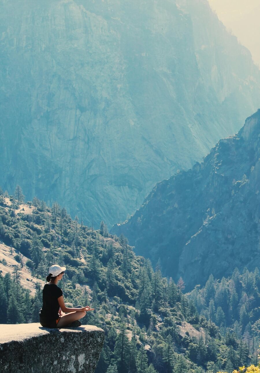 Person meditating on a cliff overlooking a forested mountain valley.